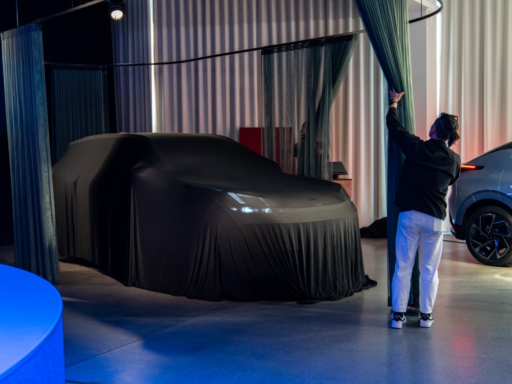 An image of a man pulling back a curtain to reveal a Lynk & Co car covered in a black sheet. The rear of another car is visible to the right. 