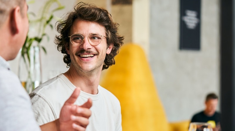 An image of a man wearing glasses in conversation with another man, who is partly visible from behind on the left side of the image. 