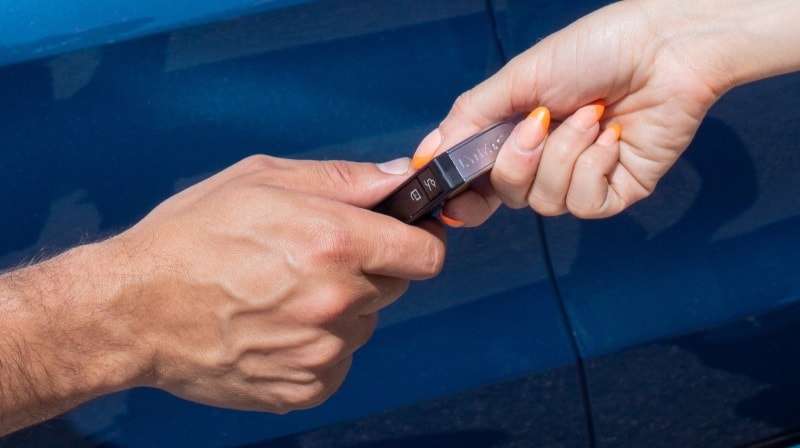 A close-up image of two hands coming together to hand over the keys to a Lynk & Co car. A panel of the car is visible in the background. 