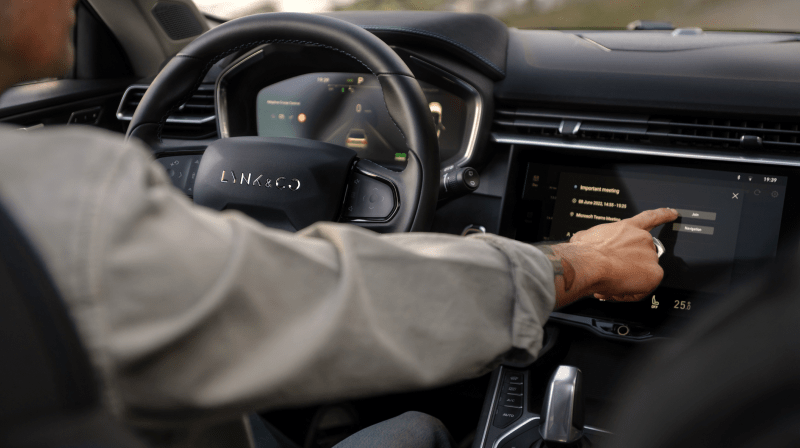 An image of a driver’s outstretched hand, interacting with something on a car’s touchscreen display. 